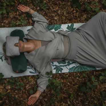 Kobieta leżąca na macie z roślinnym wzorem w lesie, z szarą poduszką na oczy i ciemnozieloną poduszką pod głową, otoczona liśćmi i naturalnym otoczeniem.